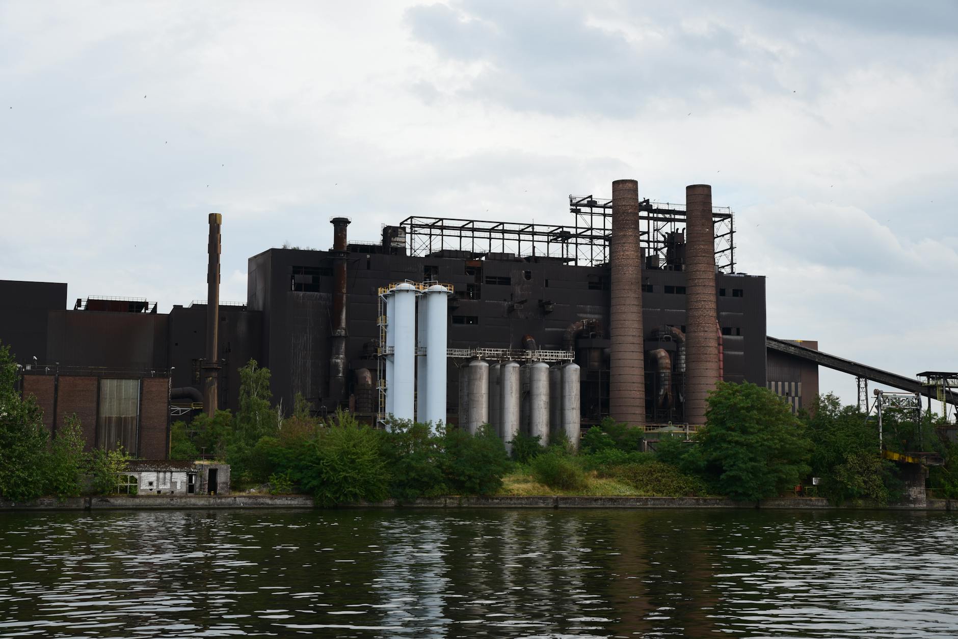 A large industrial factory with chimneys by a river, surrounded by trees on a cloudy day.