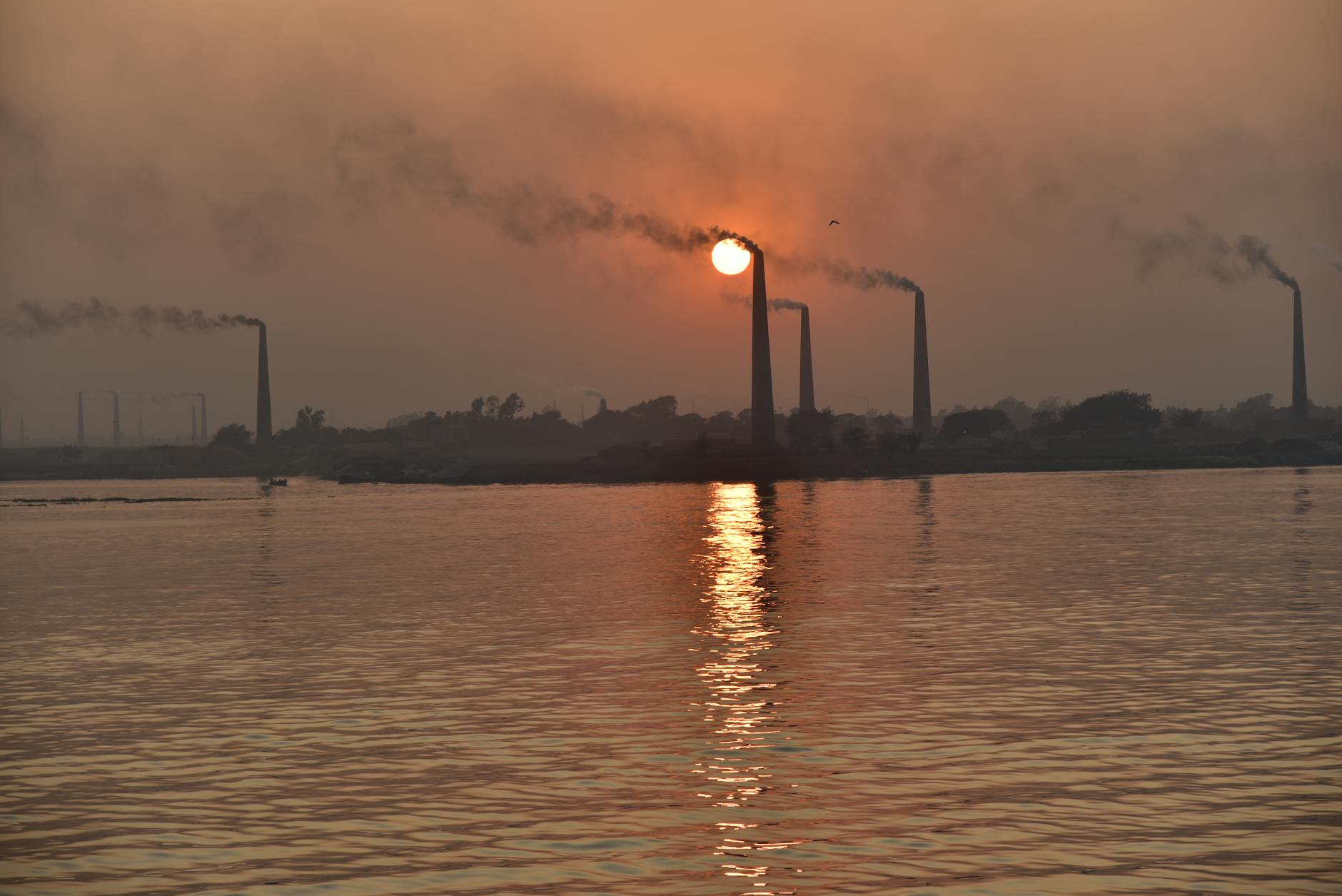 Silhouette of factory chimneys emitting smoke, reflecting on a serene lake at sunset in Bangladesh.
