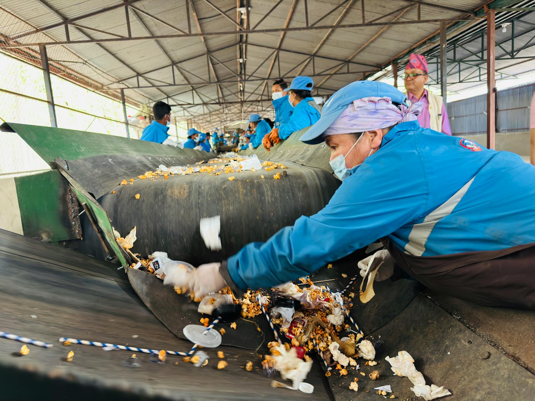 Workers in blue uniforms sorting recyclable waste on conveyor belt at a facility in Butwal, Nepal.